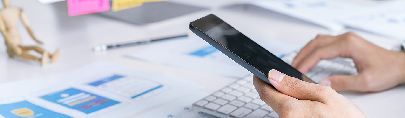 An app development professional is looking at a phone screen and typing on a keyboard simultaneously