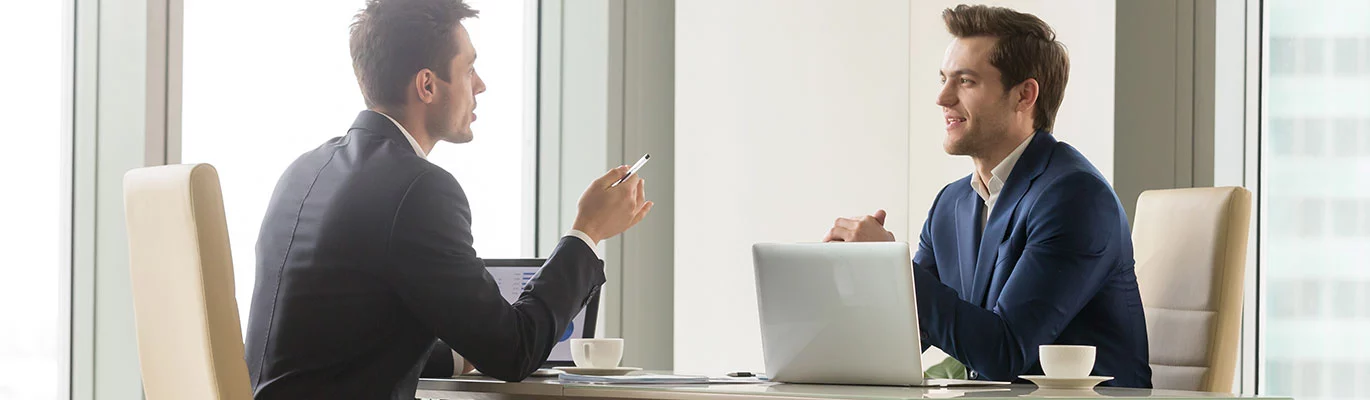 Two professionals are sitting across from each other, in front of a laptop, discussing the Microsoft Excel VBA course in Saudi Arabia and India