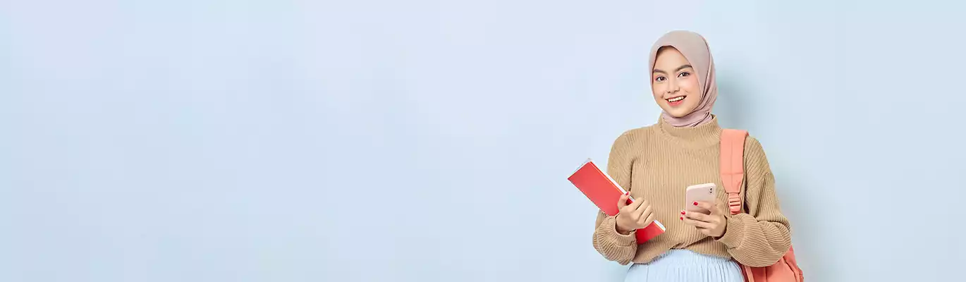 A student wearing hijab and a shoulder back standing with a mobile phone and a book in hand 