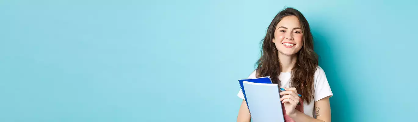 A student standing with two books in one hand and a pen in other hand, looking to the camera with a smiling face 