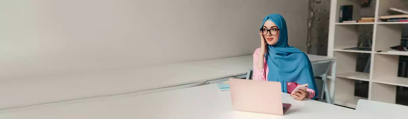 A Student is sitting infront of a laptop with a mobile phone in one hand and adjusting her specs with other hand