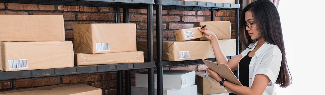 A warehouse manager standing near a shelf of warehouse goods, checking and noting down the details on it 