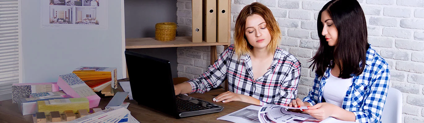 Two students with a laptop infront and a book in hand studying Interior Design Course