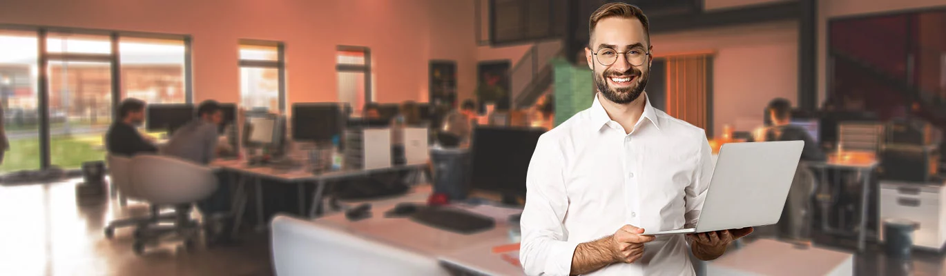 A data scientist standing with a laptop in hand in an occupied office atmosphere. He is looking to the camera with a smilin g face