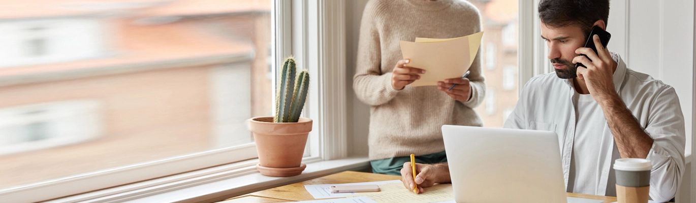 Flutter developer sitting infront of a laptop writing something while calling , another one standing with holding papers