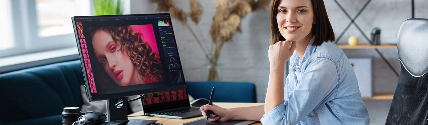 A woman holding a pen and sitting infront of a computer in a home office setting is smiling at the camera