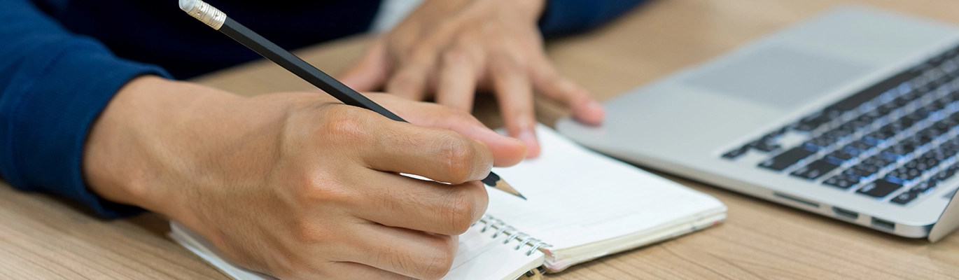 A person writing on a notepad that is sitting next to a laptop